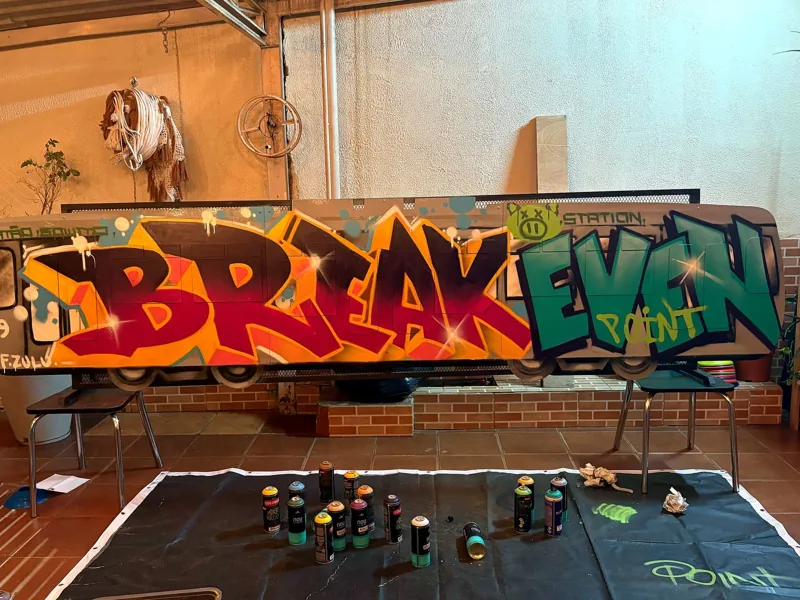 Painel pintado com as palavras Break Even Point apoiado em cadeiras, com latas de spray dispostas no chão