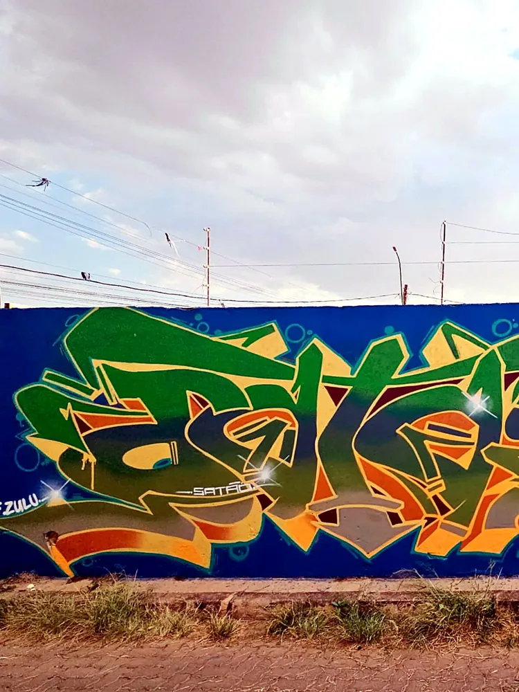 Peça de grafite verde e dourada em muro azul, campo aberto sob céu nublado