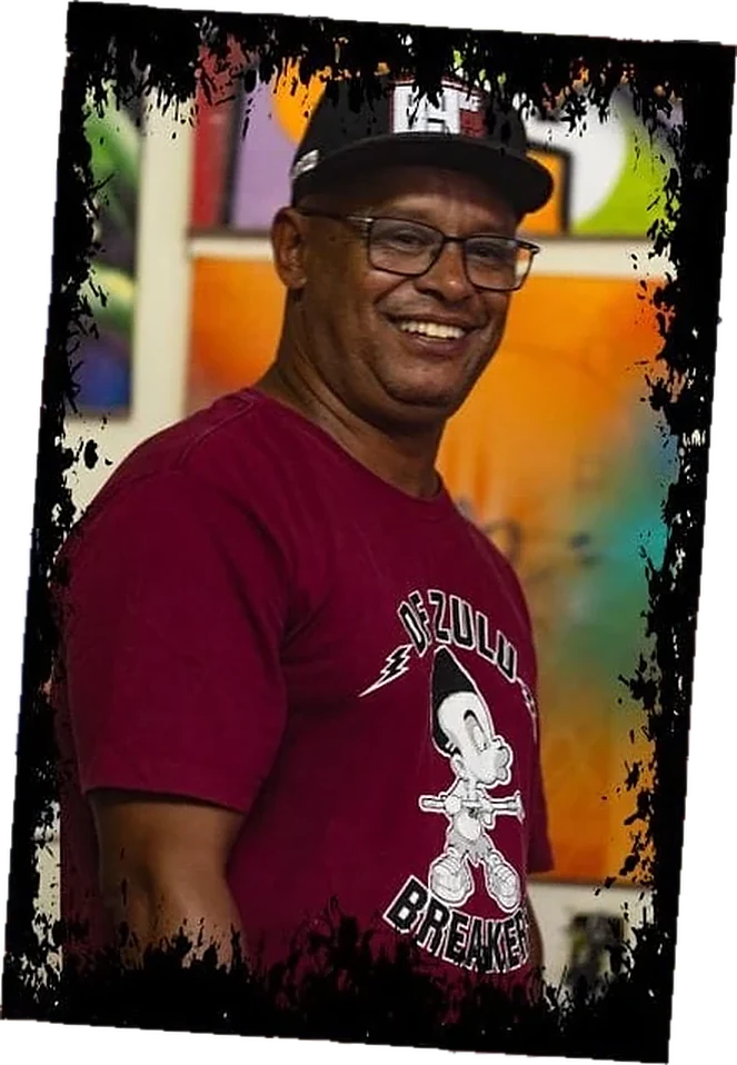 Retrato de Gilmar Satão sorrindo, de boné e óculos, vestindo camiseta vinho com estampa DF Zulu Breakers, recortado sobre a parede colorida do ateliê
